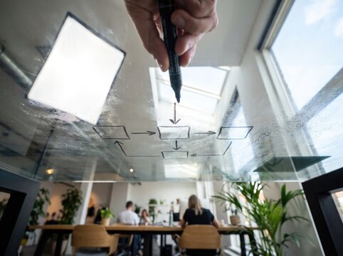 Business person hand drawing a complex flowchart diagram on a transparent glass board in a modern office studio environment with bright professional studio lighting equipment visible on the side.