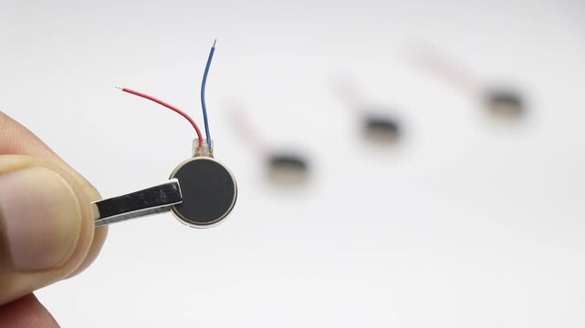Coin type micro vibration motor with red and blue wires held by tweezers with other motors blurred in background