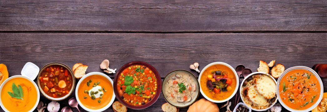 Assortment of delicious homemade warm and cozy autumn soups. Top down view bottom border on a dark wood banner background. Pumpkin, squash, sweet potato, mushroom, French onion and hearty vegetable.