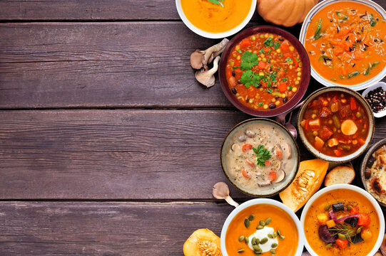 Assortment of delicious homemade warm and cozy autumn soups. Overhead view side border on a dark wood background. Pumpkin, squash, sweet potato, mushroom, French onion and hearty vegetable.