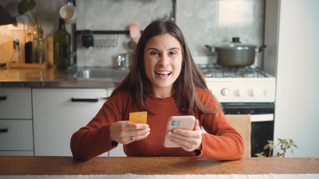 Young Caucasian woman shopping online from her home kitchen with a credit card, then looking at the camera and celebrating her purchase with excitement.
