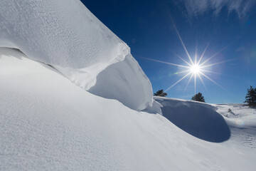 Close-up of a massive snow cornice on a mountain ridge under bright sunlight and a deep blue sky, showing fresh snow texture, dramatic shadows and pristine winter scenery. © Serhii