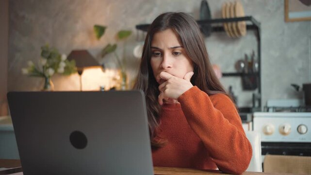 Young Latina woman using her laptop at home while looking suspiciously at the screen, highlighting online concerns, uncertainty, digital safety, and modern technology.
