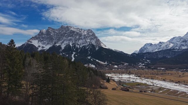 Snow-capped peaks rise above pine forests and golden fields. Clouds drift across a bright blue sky. River winds through the valley