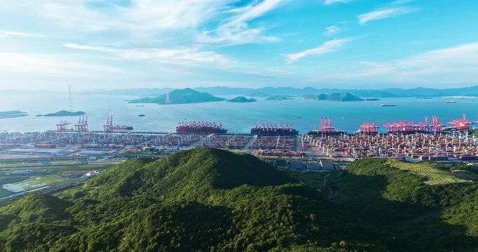 An aerial view of a busy container port terminal and coastline framed by lush green hills under bright sunlight and a blue sky with clouds.