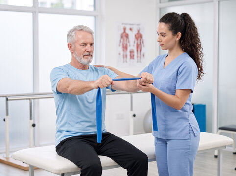 young female physiotherapist assisting senior man during seated band assisted shoulder flexion exercise in clinic
