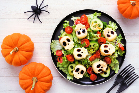 Spooky Halloween skull salad. Above view table scene on a white wood background. Healthy greens and vegetables with creepy cheese skull heads.