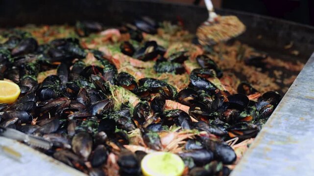 Chef cooking large pan of seafood paella with mussels, shrimp and lemon. Street food preparation at market. Traditional spanish rice dish with shellfish
