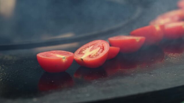 Halved red tomatoes cooking on hot black griddle with rising smoke. Fresh vegetables roasting on pan, healthy food preparation, street food and restaurant cuisine concept.