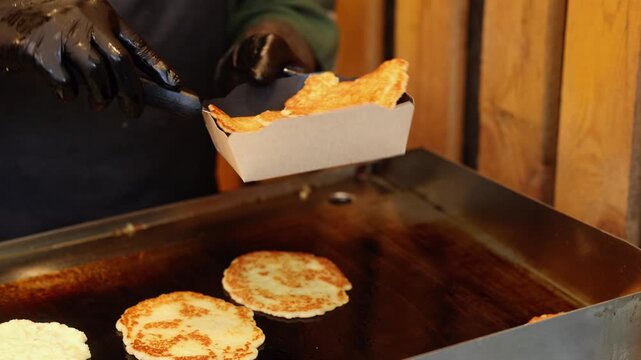 Man in black gloves cooking pancakes on griddle and packing them into paper box. Street food vendor preparing fresh pancakes for takeout order