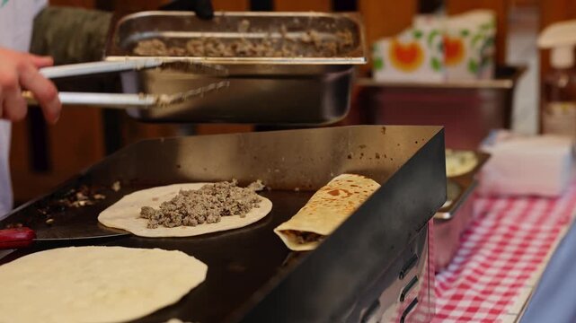 Chef cooking folded tortilla wraps with meat filling on flat grill at street food stall. Cook in black gloves preparing quesadilla with spatula. Fast food preparation concept