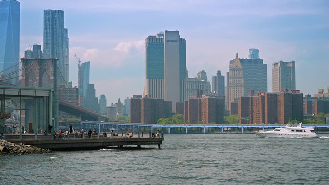 New York, USA, 1 May 2026 : Motor yacht cruising past Manhattan skyline and Brooklyn Bridge. People watch from a waterfront pier under a hazy blue sky in New York.