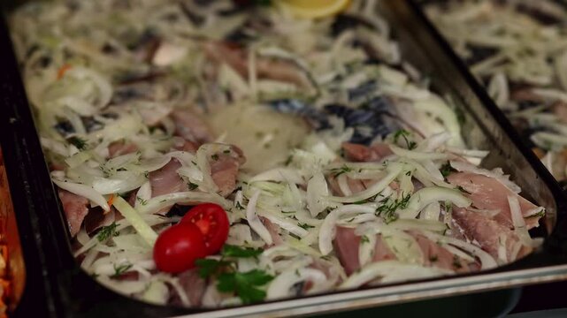 Marinated herring pieces with onion rings and spices in metal tray. Pickled fish appetizer with tomato and herbs on buffet counter. Traditional seafood snack food closeup