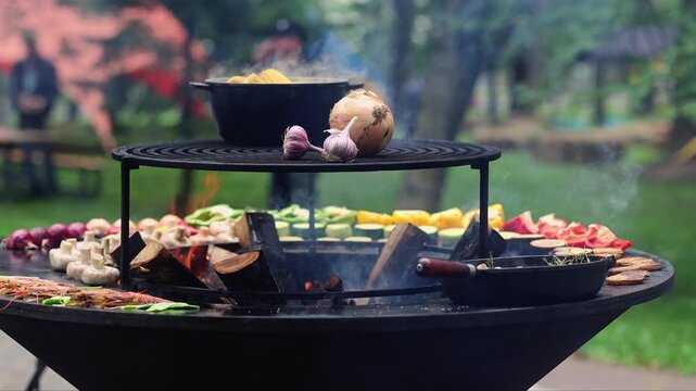 Vegetables and garlic cooking on round open fire grill in park. Outdoor barbecue with flame and smoke. Summer picnic food preparation on charcoal brazier concept