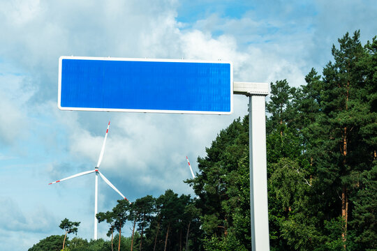 Blank blue motorway sign on a pole with wind turbines and green trees under a cloudy sky, concept of road signage mockup with copy space