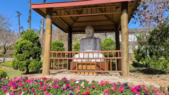 Great Buddha monument replica of the famous historical Daibutsu of Kamakura in the Kanagawa prefecture of Japan. Traditional wooden pavilion,  vibrant pink and purple flowers. Campos do Jord&atilde;o.