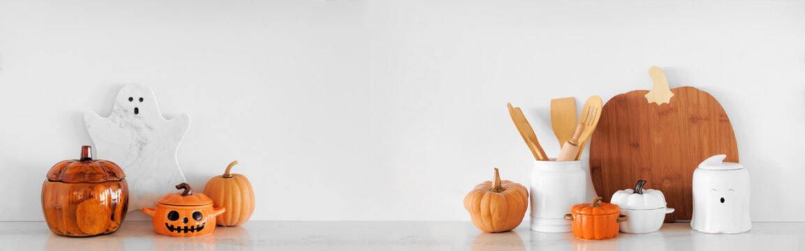 Halloween ghost and pumpkin theme kitchenware on a white stone countertop. Banner against a white wall background with copy space. Autumn kitchen decor concept.