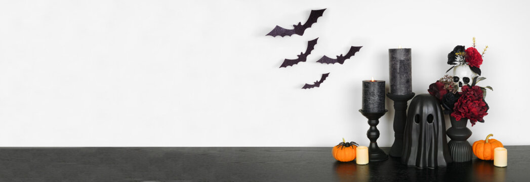 Halloween gothic shelf decor with copy space against a white wall banner background. Black shelf with glowing candles, dark flowers, skull, pumpkins, bats, ghost and spider.