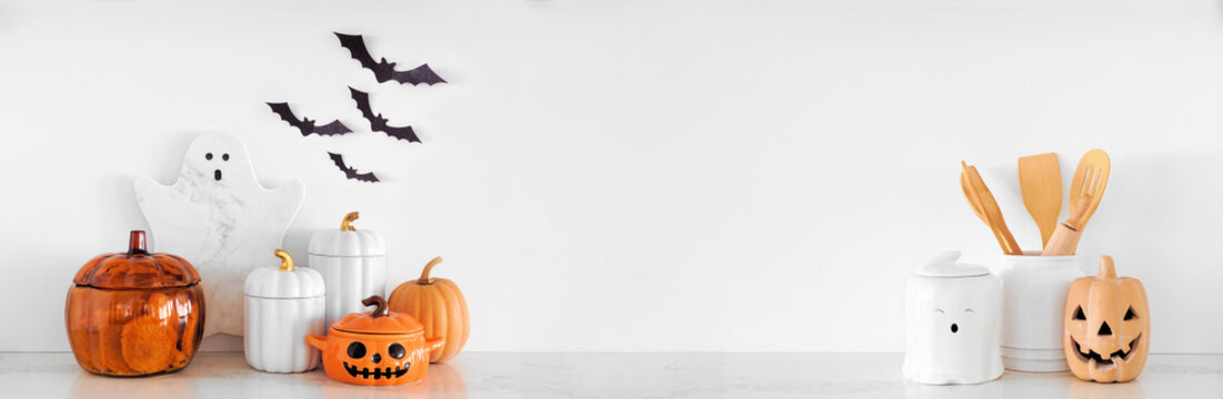 Halloween ghost and pumpkin theme kitchenware on a white stone countertop. Banner against a white wall background with copy space. Autumn kitchen decor concept.