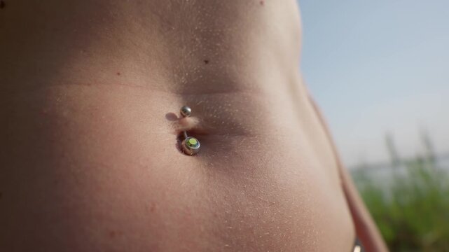 A close-up view shows a silver belly button piercing on a woman stomach. Warm sunlight illuminates natural skin texture in the outdoors. The sparkling crystal reflects the summer light.