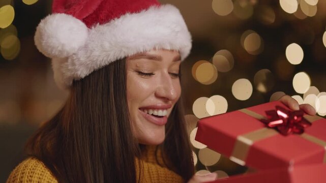 Curious woman in Santa hat opening wrapped present, looking inside gift box with pleasant surprise and excitement
