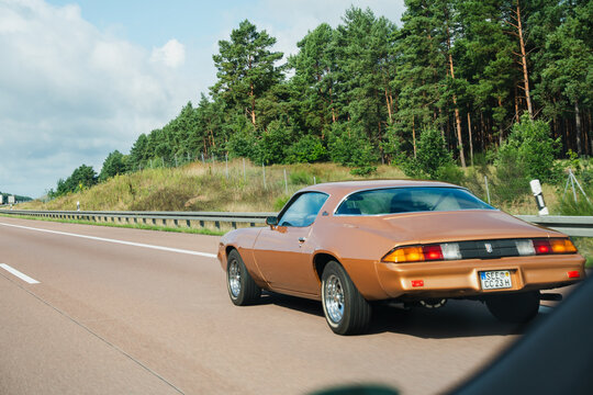 Germany - Aug 22, 2026: Rear view of a brown classic muscle car with round tail lights cruising along the A12 autobahn beside a grass embankment and pine forest under scattered clouds