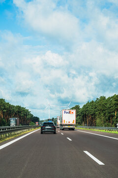 Germany - Aug 22, 2026: FedEx delivery truck and a dark car driving on the A12 autobahn with wide asphalt in the foreground, roadside pines and heavy white clouds above the carriageway