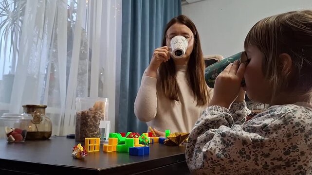 Mom drinks coffee while daughter plays with blocks in a room with natural light