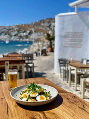 Na drewnianym stoliku nadmorskiej restauracji stoi talerz z sałatką z jajek, ziemniaków i ziół oraz szklanka zimnego piwa. W tle rozciąga się morze i malownicze miasteczko.