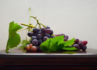 Classic still life composition featuring a fresh bunch of ripe purple and red grapes with vibrant green vine leaves and stems, resting on a polished dark wooden surface against a wall background. © Alena