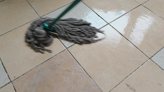 Gray string mop cleans across tiled flooring in a wiping motion, floor cleaning routine, household maintenance scene.