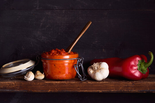 Homemade ajvar made from roasted red peppers and garlic in a glass jar, with fresh red pepper and garlic on a rustic wooden shelf.