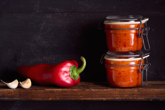 Homemade ajvar made from roasted red peppers and garlic in a glass jar, with fresh red pepper and garlic on a rustic wooden shelf.