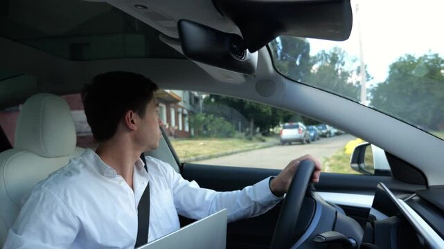 Young manager driving modern electric car reversing out of his home driveway. Male entrepreneur keeps close eye on the mirrors and rearview camera ensuring safe start to morning commute to the office