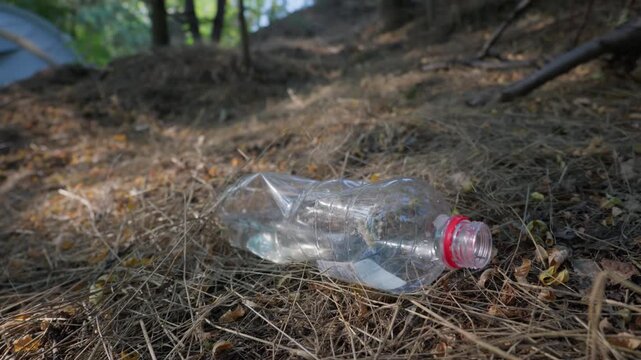 An empty plastic bottle lies discarded on dry grass in a campground forest near a tent. Concept of irresponsible camping, environmental pollution, plastic waste, and ecotourism impact.
