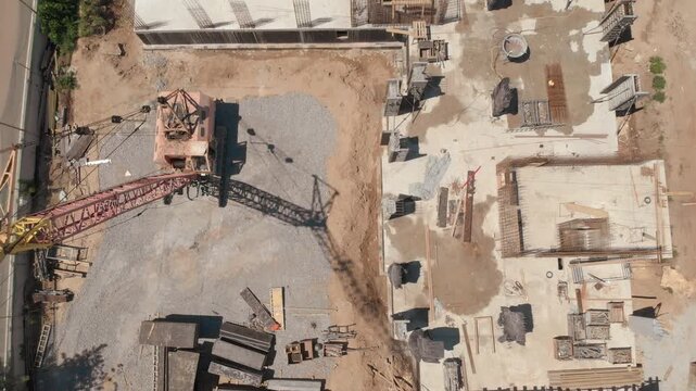 Top-down aerial view of tower crane above gravel staging area beside concrete foundation with rebar cages, formwork panels, scaffolding, and timber boards under clear daylight