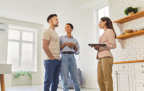 Real estate agent shows a young couple an apartment, pointing out the bright kitchen while reviewing purchase steps. Woman realtor showing new house to clients going to buy a property. 