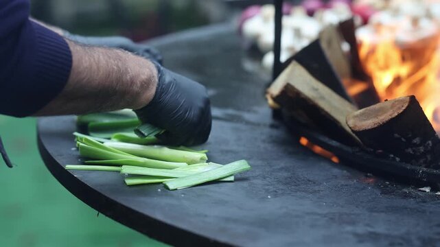 Man in black gloves grilling green leeks on round fire grill with burning wood. Chef cooking vegetables over open flame. Outdoor barbecue and fire cooking concept.