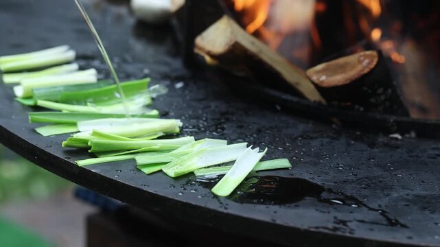 Green leek pieces grilling on hot outdoor stove with open fire flame. Oil poured onto fresh vegetables on griddle. Outdoor cooking, barbecue and healthy food preparation concept.