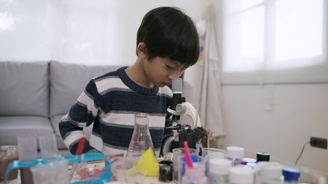 Happy adorable Asian boy with microscope sitting learning science and conducting chemistry experiments by mixing various colored liquids with test tubes and beakers, enjoying in nursery classroom lab.
