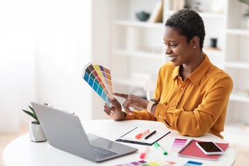 Smiling Black Woman Engaged In Design Work, Pointing At Color Palette While Sitting At Modern Home...