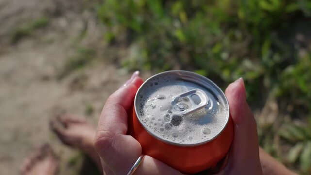 Hands hold an open aluminum can with fizzy carbonated foam rising at the beach in slow motion. Bright daylight illuminates the refreshing drink during a relaxing summer picnic.