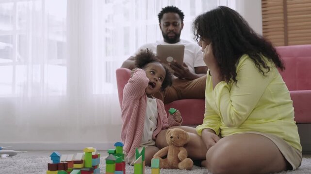 Family day concept. Happy African family is depicted with a mother and a cute little daughter playing with wooden blocks on the floor while the father sits on the sofa in the living room using tablet.