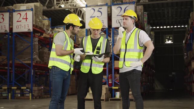 Teamwork of three warehouse workers in uniform wearing yellow safety helmets, standing and working talking and discussing planning in large logistics distribution factory. Inventory stock concept. 