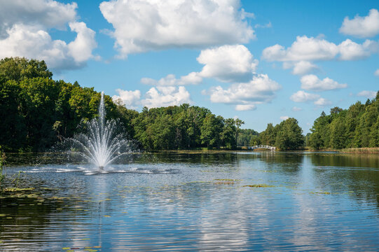 Lake with a central water fountain surrounded by green trees and a small white bridge in the distance