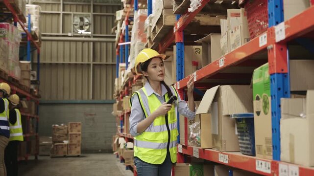 Team of Asian warehouse workers in uniform with yellow safety helmets holding and using a barcode scanner for checking inventory stock on shelves together in a logistics distribution factory center. 