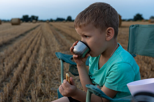 Young boy drinking from a reusable metal cup and eating a snack while resting in a harvested field. Concept of outdoor picnic, healthy childhood, hydration, sustainable lifestyle and rural adventure.