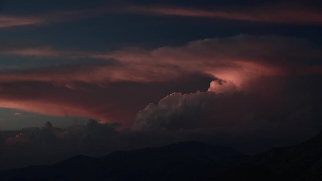 Sunset glows behind a distant thunderstorm, coloring cloudbank and hills with warm dusk light. Dark sky and clearing storm create a dramatic weather scene with orange tones and mountain silhouette.