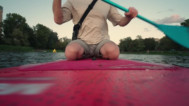 Man Paddling Stand-Up Paddleboard on Urban River at Dusk: Low-Angle View from Board | Clean Water City Recreation Kayaking Lifestyle | Sustainable Green Urban Living