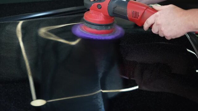 Close up of a professional car detailing process. Mans hands hold a red electric orbital dual action polisher machine with a purple microfiber wool pad buffer tool to polish a black auto paint hood.